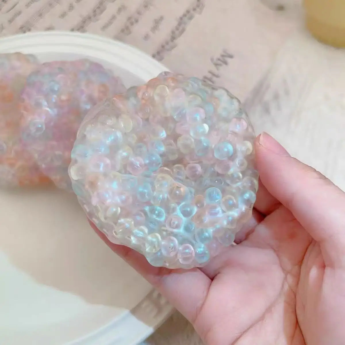 A hand holds a clear, iridescent squishy stress relief ball with blue and pink hues.