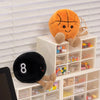 An orange plush sports ball companion with feet sits next to a white storage box filled with various small items.