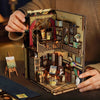 A hand holds an open DIY wooden book nook kit featuring miniature figurines and detailed architectural elements.