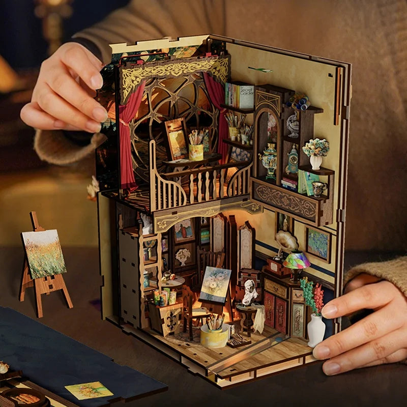 A hand holds an open DIY wooden book nook kit featuring miniature figurines and detailed architectural elements.