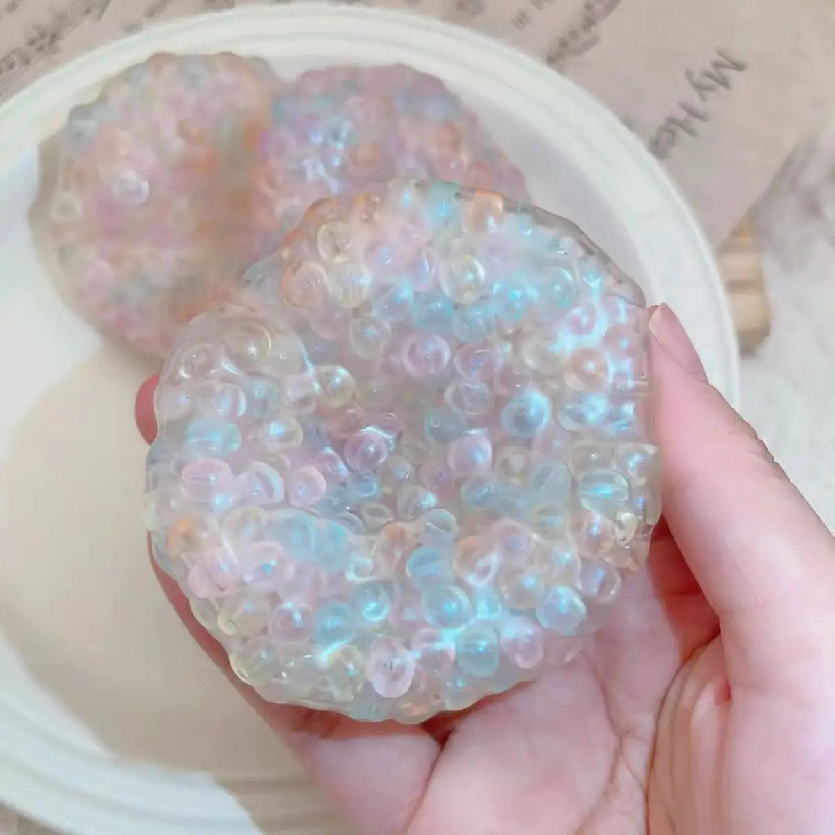 A hand holds a clear, iridescent squishy stress relief ball with pink and blue hues.