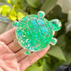 A hand holds a translucent green squishy star turtle fidget ball with sparkling blue and green glitter.