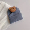 A knitted baby hat in gray with a brown teddy bear ear decoration resting on a white ribbed fabric.