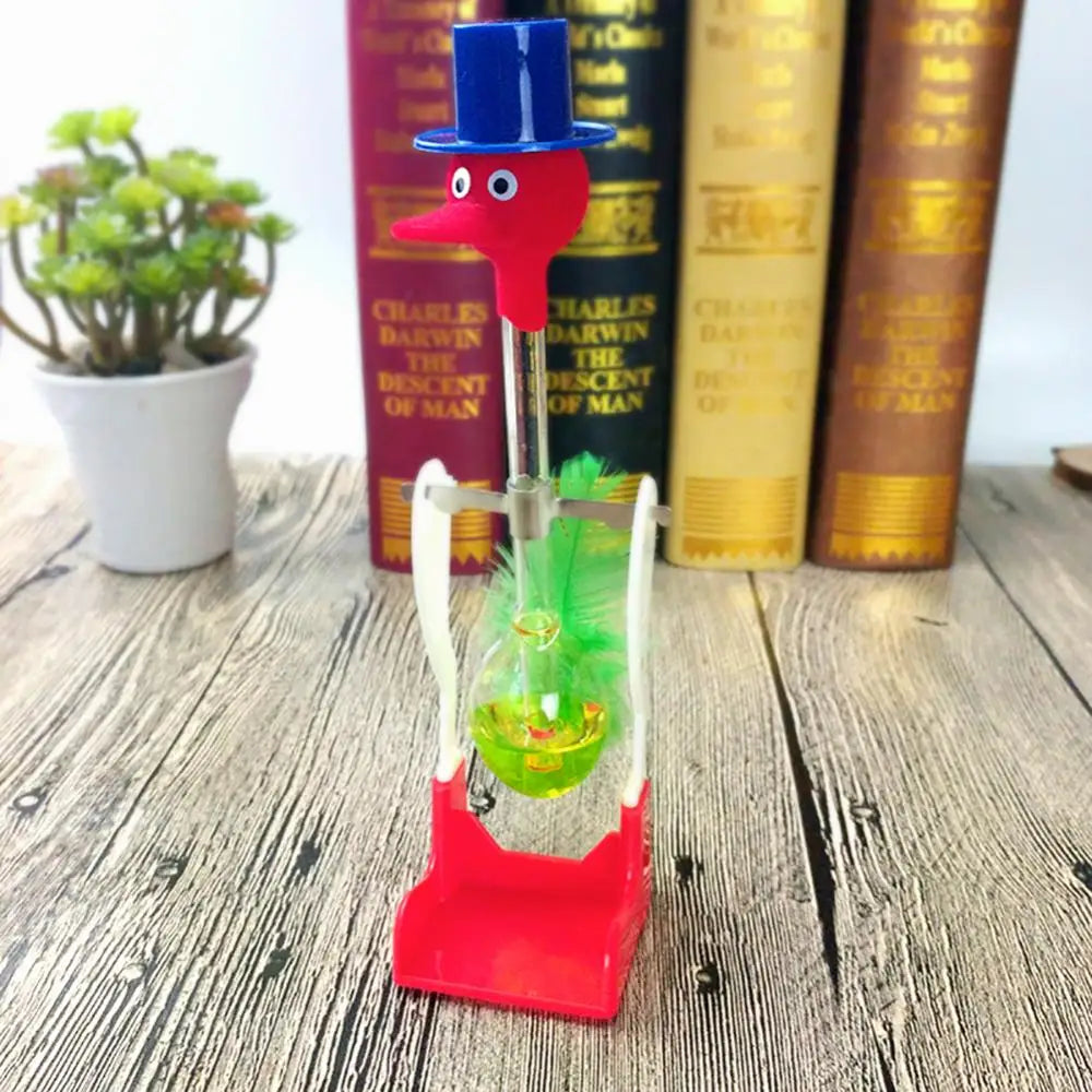 A red and blue drinking bird toy with a green feather is displayed in front of books on a wooden surface.