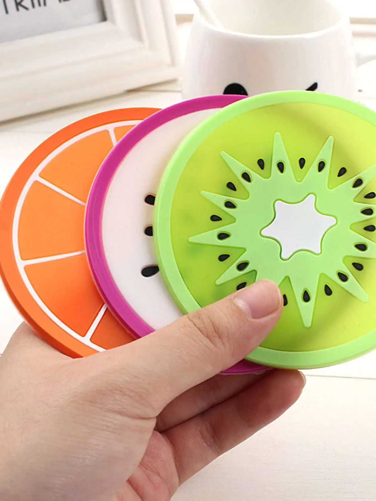 A hand holds a set of colorful fruit pattern PVC coasters, including orange and lime green designs.