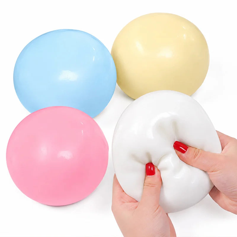 A hand holds a white jumbo slow-rising cheese squishy stress relief toy next to three colored balloons: blue, yellow, and pin