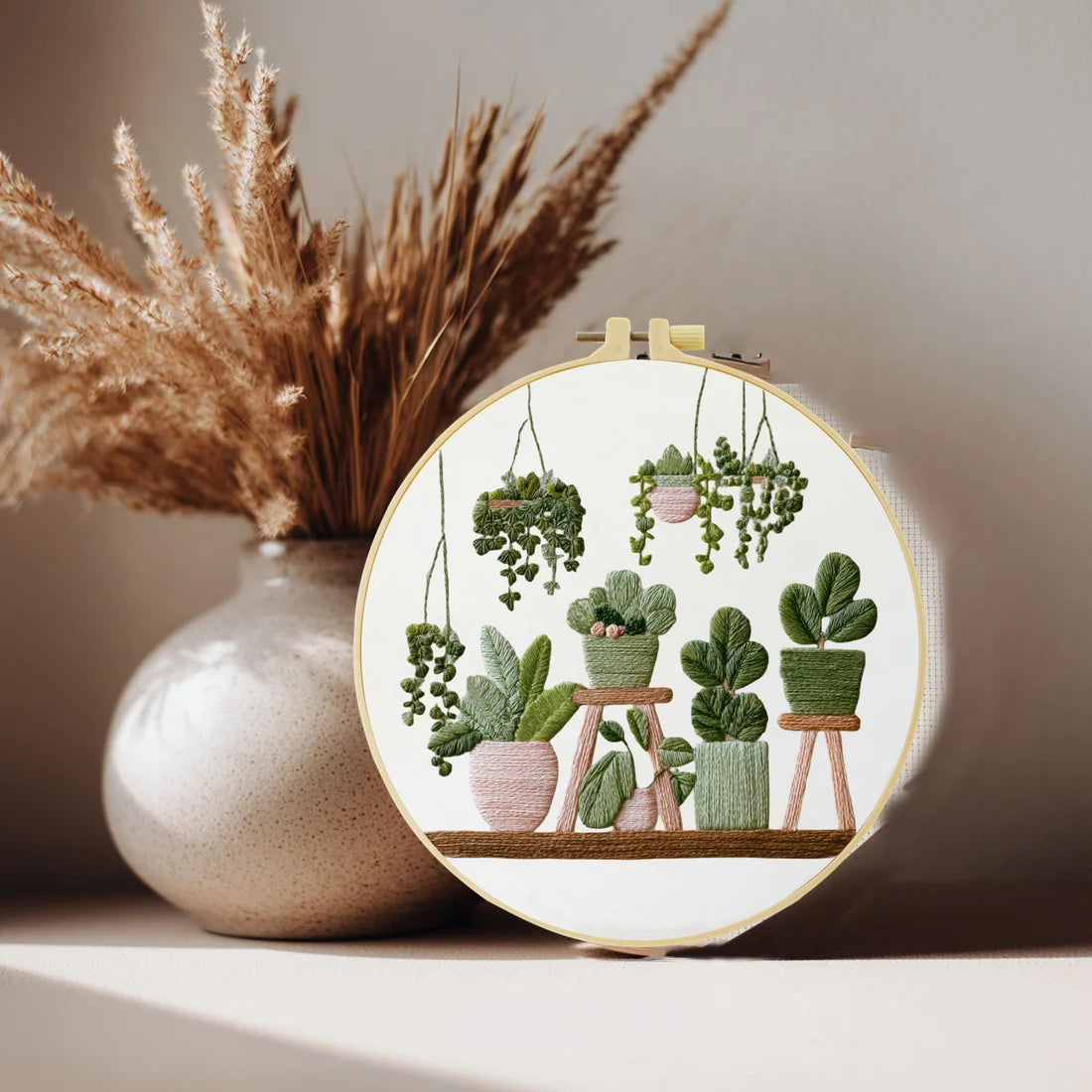 A circular embroidery hoop featuring various green potted plants and foliage on a wooden stand.