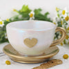 Une tasse en céramique blanche avec une anse dorée repose sur une soucoupe, avec un moule en silicone en forme de cœur à prox