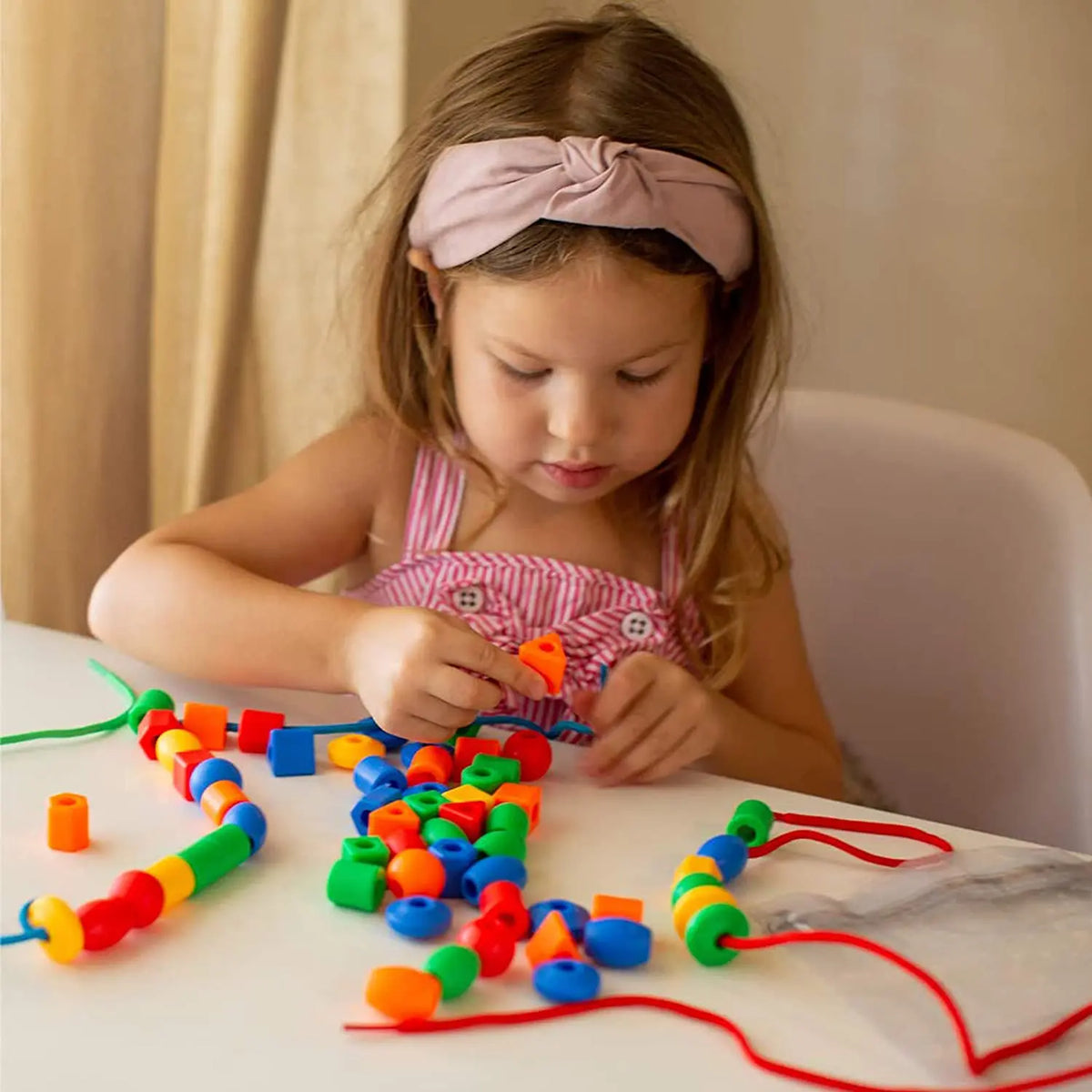 A young girl is threading colorful rainbow lacing beads onto strings on a white table.
