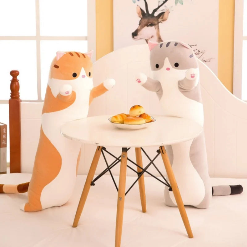 A plush cat pillow resembling a tiger is sitting next to a gray cat pillow on a white table.