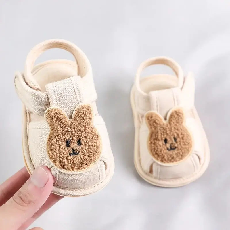 A pair of beige soft sole sandals for baby's first steps featuring a cute bear face design.