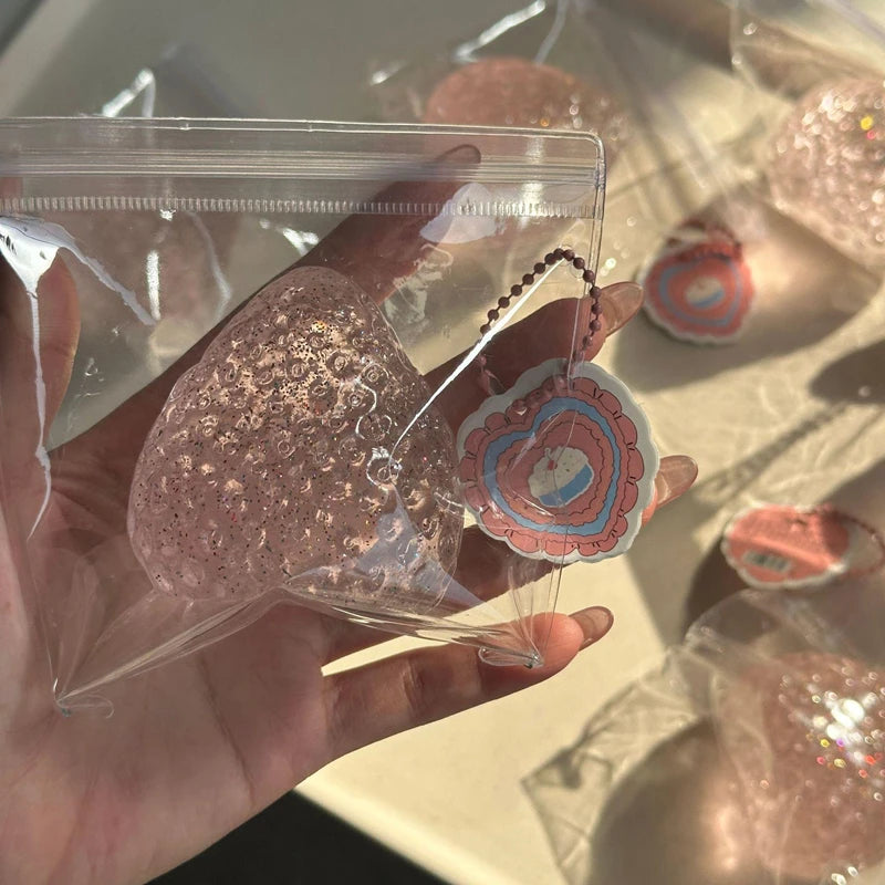 A hand holds a clear plastic bag containing a pink strawberry squeeze fidget toy.
