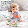 A baby is playing with a Montessori Magic Tissue Box Ocean cloth book and a small green dinosaur toy on a white blanket.