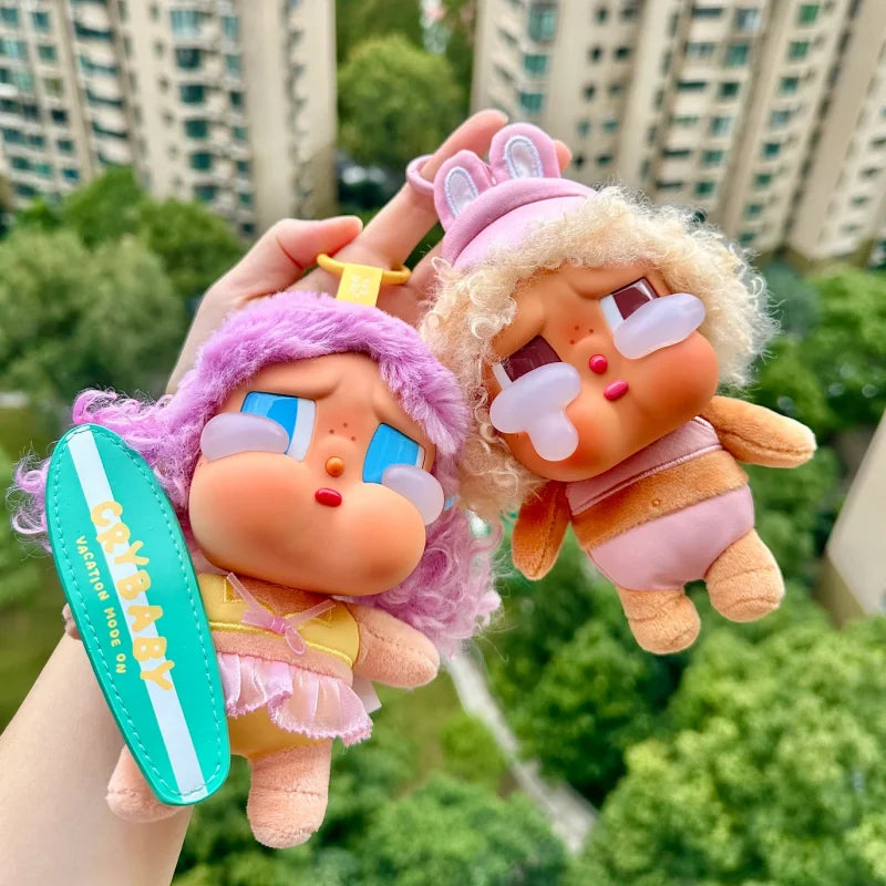 A hand holds a Crybaby Vacation Mode On vinyl figure blind box with pink hair and light purple clothing.
