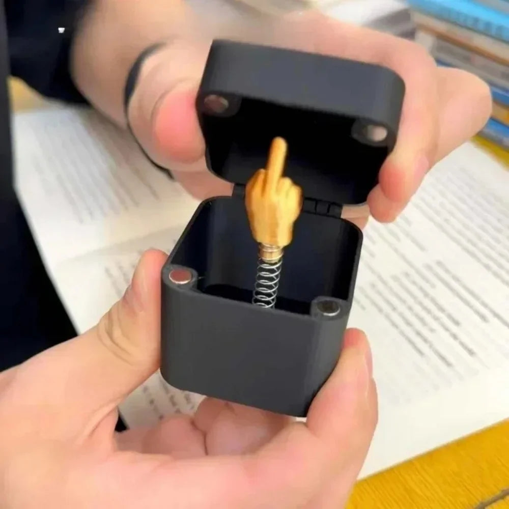 A hand is opening a black gift box containing a golden middle finger prank item.