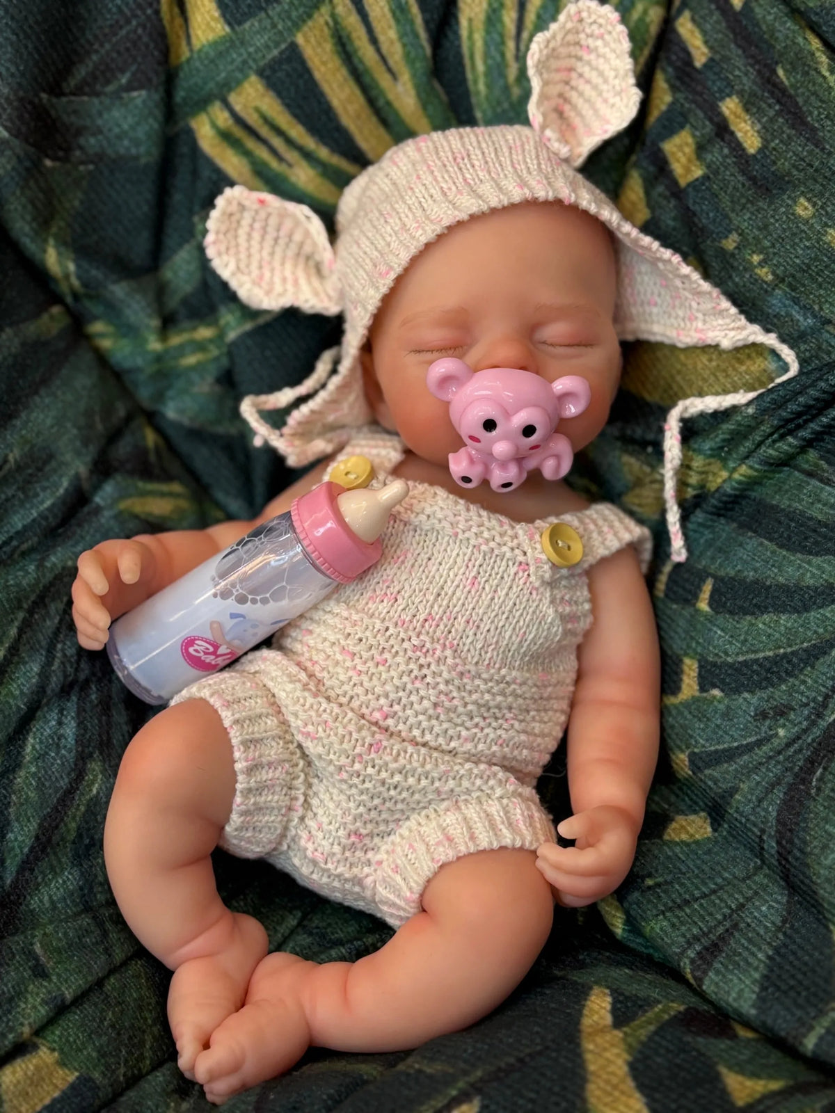 A hand-painted silicone reborn doll with pink ears and a light pink knitted outfit lying on green fabric.