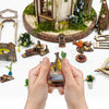 A hand holds a miniature wooden floral book nook kit with an LED light, set against a backdrop of moss and miniature greenery