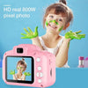 A young girl is smiling while applying green face paint to her face, with a pink mini digital camera in the foreground.