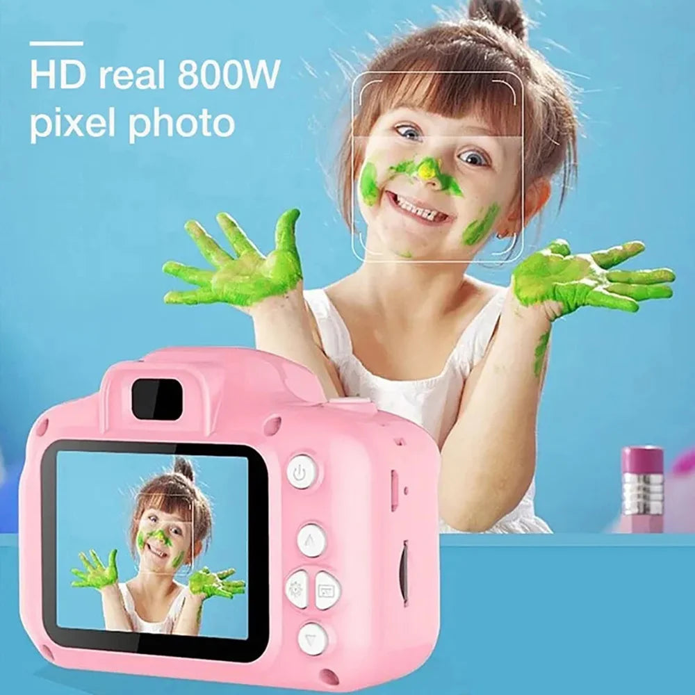 A young girl is smiling while applying green face paint to her face, with a pink mini digital camera in the foreground.