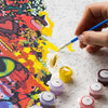 A hand paints a DIY paint by number kit featuring a seaside flower window scene on canvas with various colored paint pots.