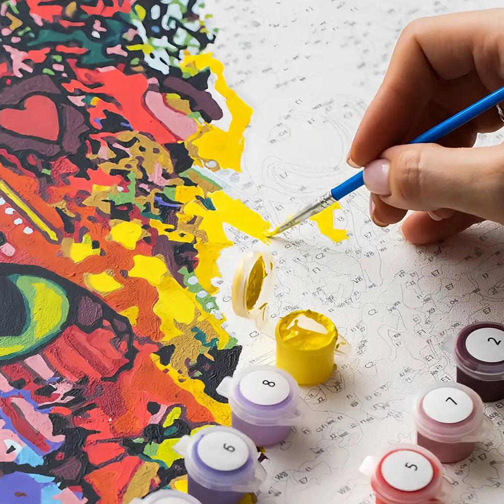 A hand paints a DIY paint by number kit featuring a seaside flower window scene on canvas with various colored paint pots.