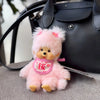 A fluffy pink cartoon bear plush toy resting on a gray surface next to a black handbag.
