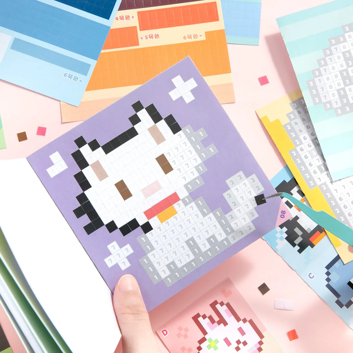 A hand holds a purple square sticker sheet featuring a black and white cartoon animal design, surrounded by colorful paper an