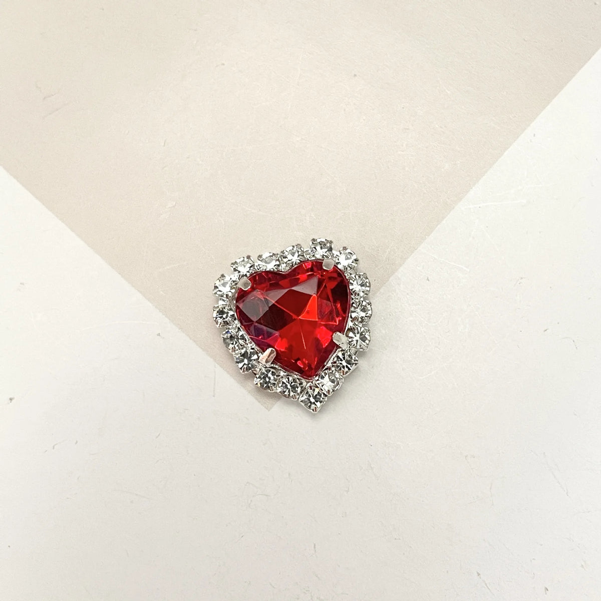 A brilliant red heart-shaped gemstone surrounded by clear rhinestones for DIY crafts.
