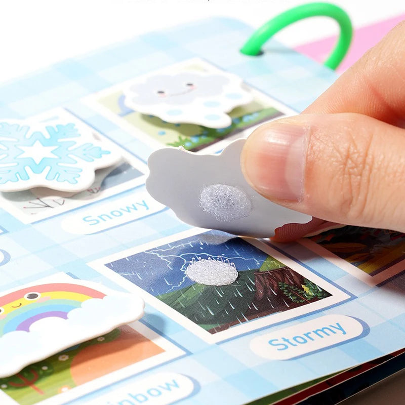 A finger is placing a gray cat sticker onto an interactive learning book featuring various weather-themed stickers like snowy