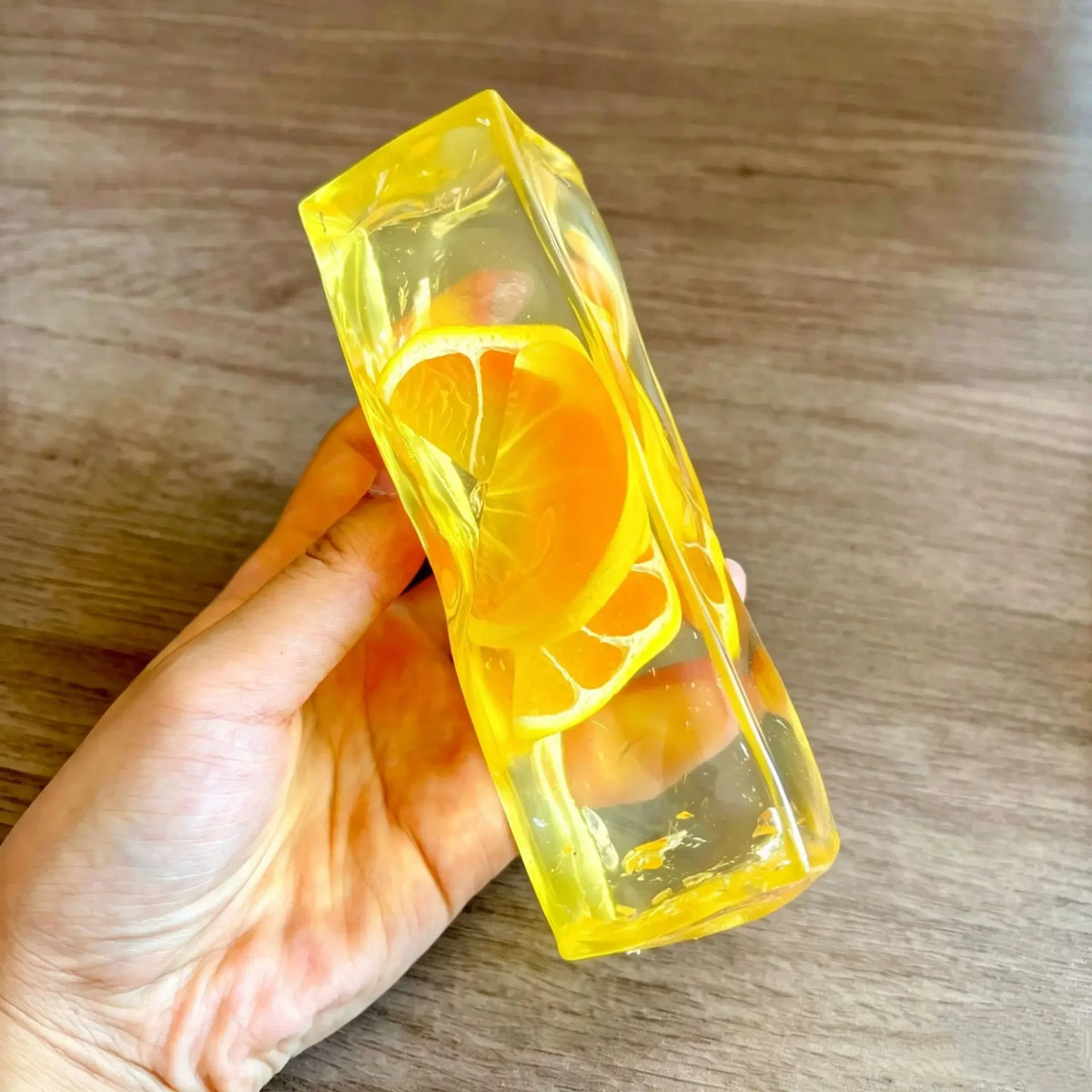 A hand holds a clear yellow ice cube shaped like a slice of orange on a wooden surface.