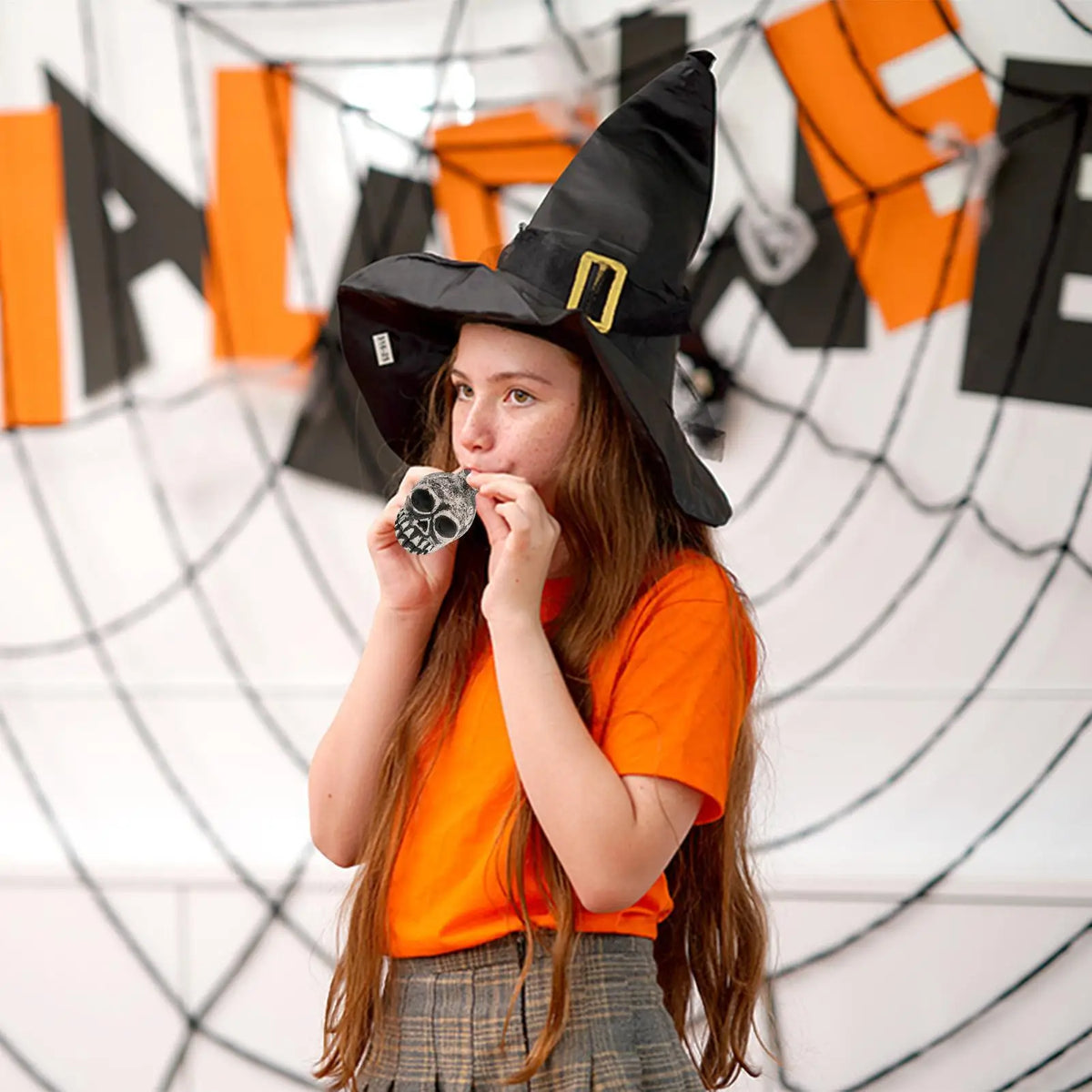 A young woman wearing a witch hat and orange shirt is holding a ceramic Aztec skull whistle.