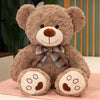 A brown teddy bear plush pillow with a plaid bow resting on a pink surface.