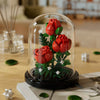 A glass dome display of a buildable eternal rose flower kit featuring vibrant red roses and green foliage.