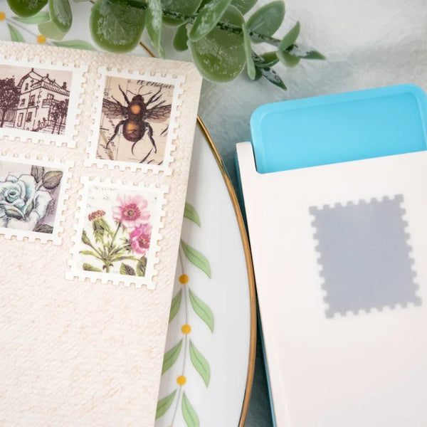 A white crafting pad features several postage stamps, including a bee and a rose, next to a light blue punch tool.