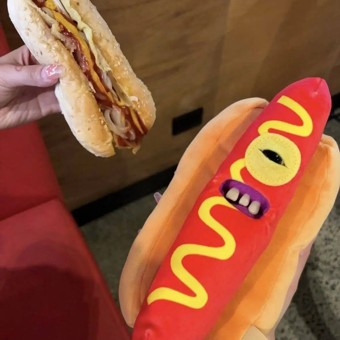 A hand holds a sandwich and a plush toy of an ugly cute monster in red and orange colors.