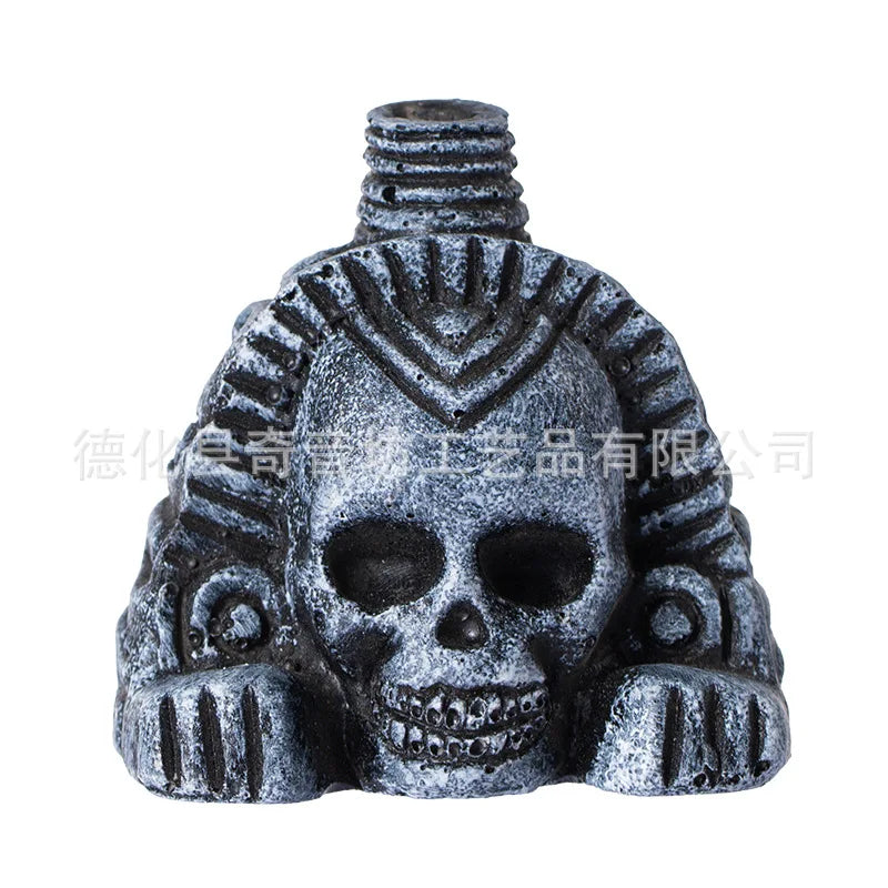 A dark gray skull with intricate patterns and facial features, resembling an Aztec death whistle or screaming noise maker.