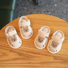 A pair of light beige soft sole first walker shoes for babies with bear-shaped fabric accents resting on a wooden surface.