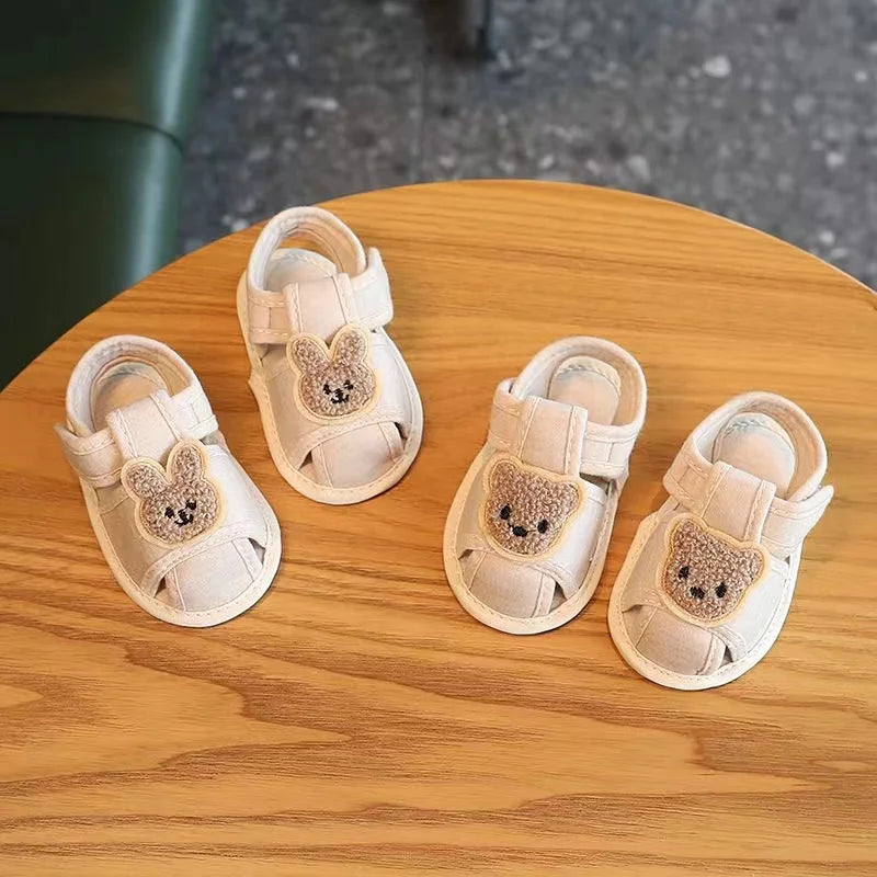 A pair of light beige soft sole first walker shoes for babies with bear-shaped fabric accents resting on a wooden surface.