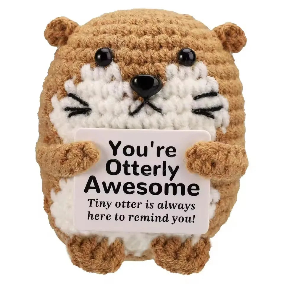 A handmade crochet owl toy with brown fur and white facial markings, featuring a sign that reads You're Otterly Awesome.
