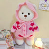 A pink teddy bear plush wearing a matching pink dress with red hearts, displayed on a white surface.