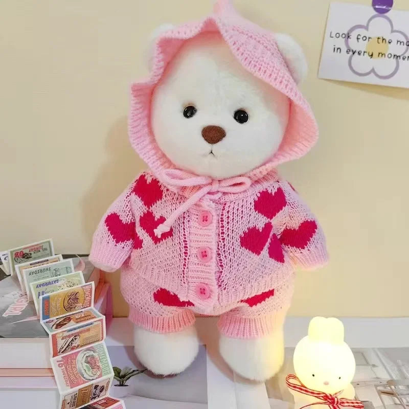 A pink teddy bear plush wearing a matching pink dress with red hearts, displayed on a white surface.