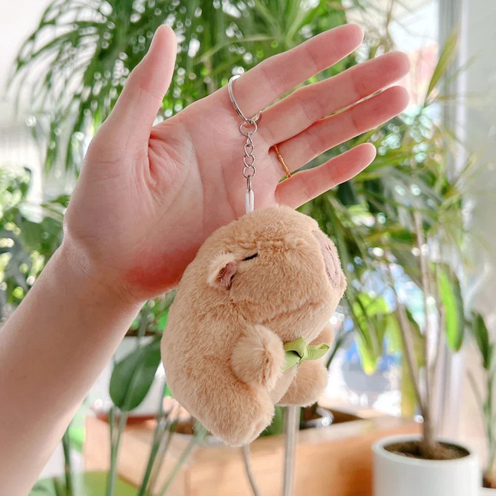 A hand holds a soft brown capybara plush keychain charm in front of a leafy indoor plant.