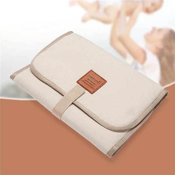 A beige foldable waterproof baby changing pad with a brown strap is shown on a wooden surface.