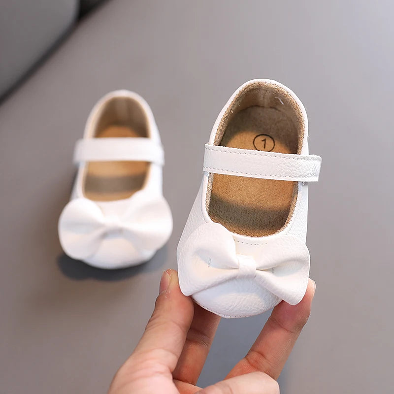 A hand holds a white lace-trimmed baby girl moccasin shoe with a bow, showing the interior and exterior detail.