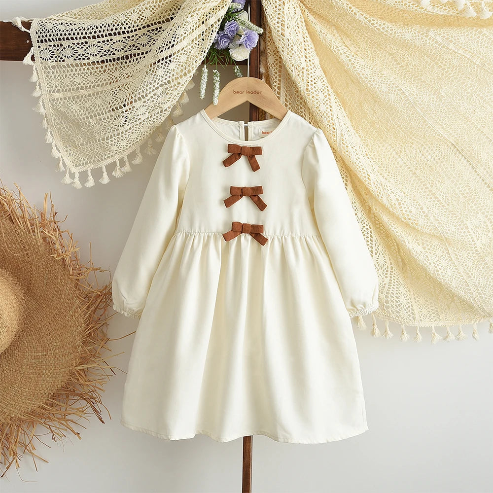 A cream-colored girls' long sleeve bow detail A-line dress hanging against a light background.