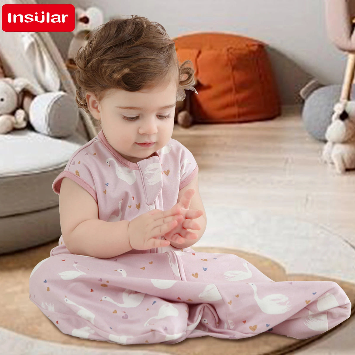A baby wearing a pink cotton sleeping bag sleeve with white swan patterns is sitting on the floor.