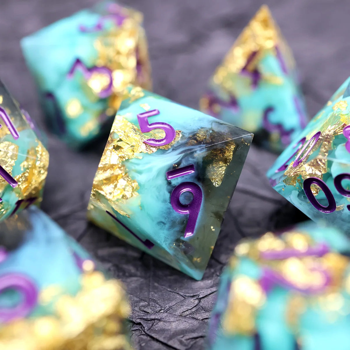 A close-up of several teal resin polyhedral dice with gold accents scattered on a dark surface.