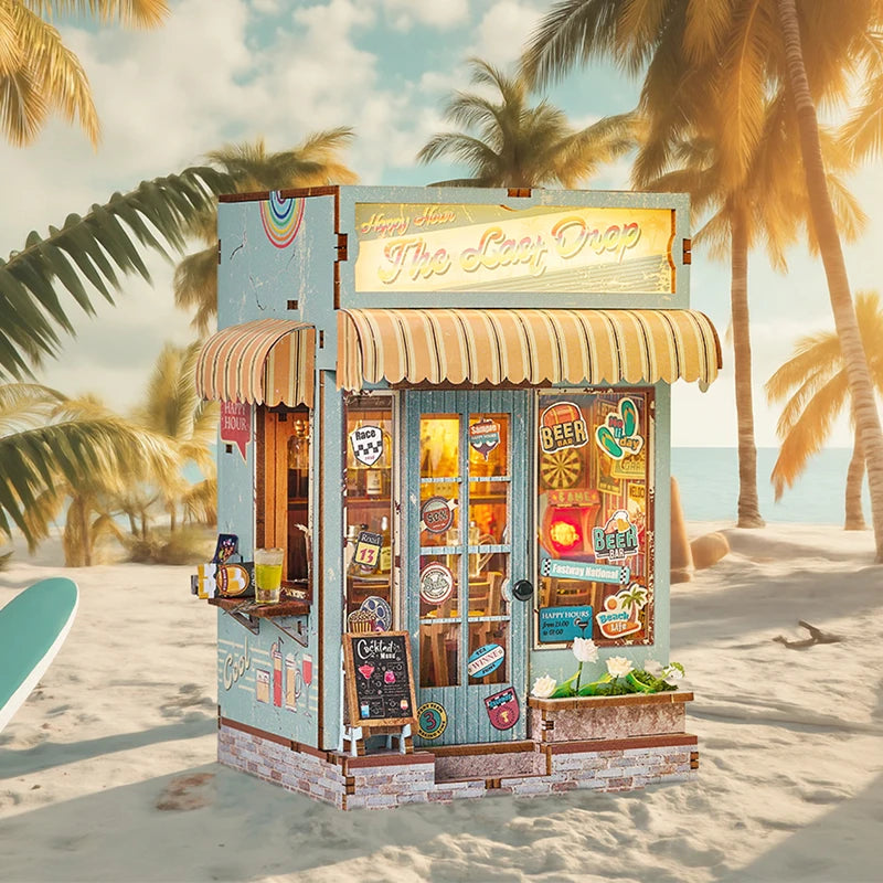 A miniature wooden book nook kit with a striped awning and glass display for dollhouse decor on a sandy beach.