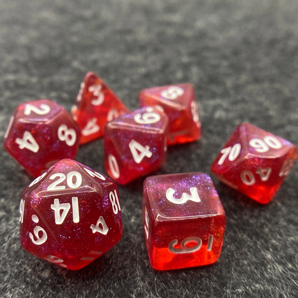 A collection of seven iridescent red acrylic polyhedral dice scattered on a dark surface.