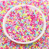 A white bowl filled with colorful polymer clay slice sprinkles for slime and DIY crafts scattered on a pink surface.